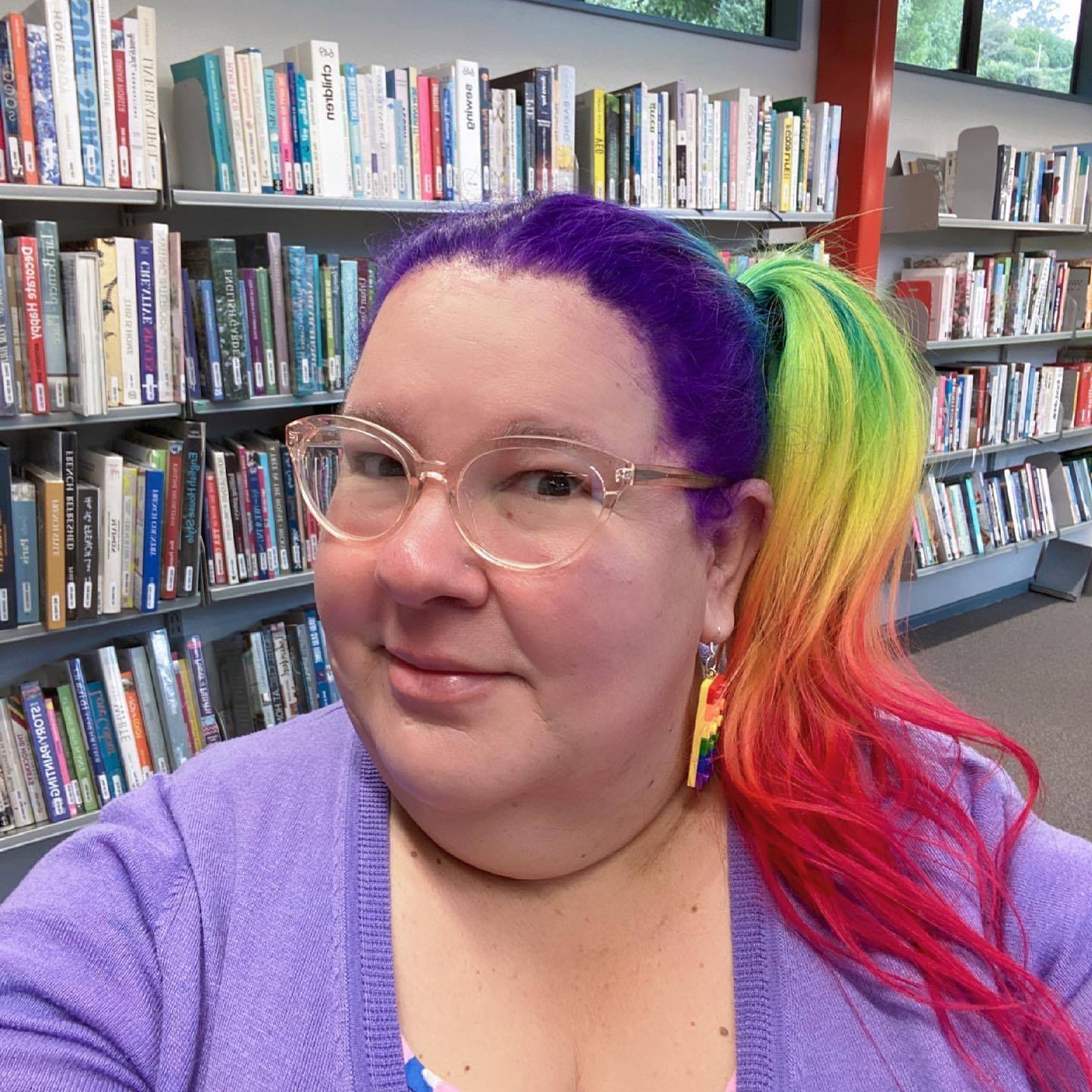 A selfie of Kath - a fat babe - in a library. Kath wears glasses and has rainbow coloured hair